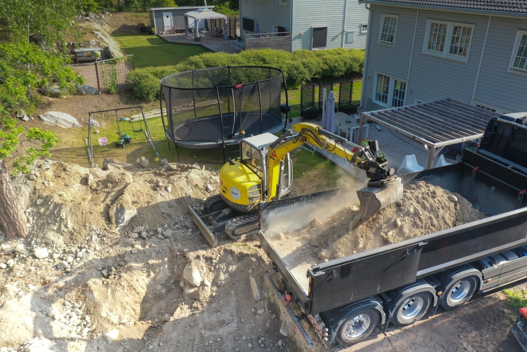 Grävmaskin utför schaktning bredvid villa och fyller lastbilsflak med utgrävd jord