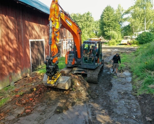 Röd grävmaskin förbereder dikesgrävning bredvid en ladugård